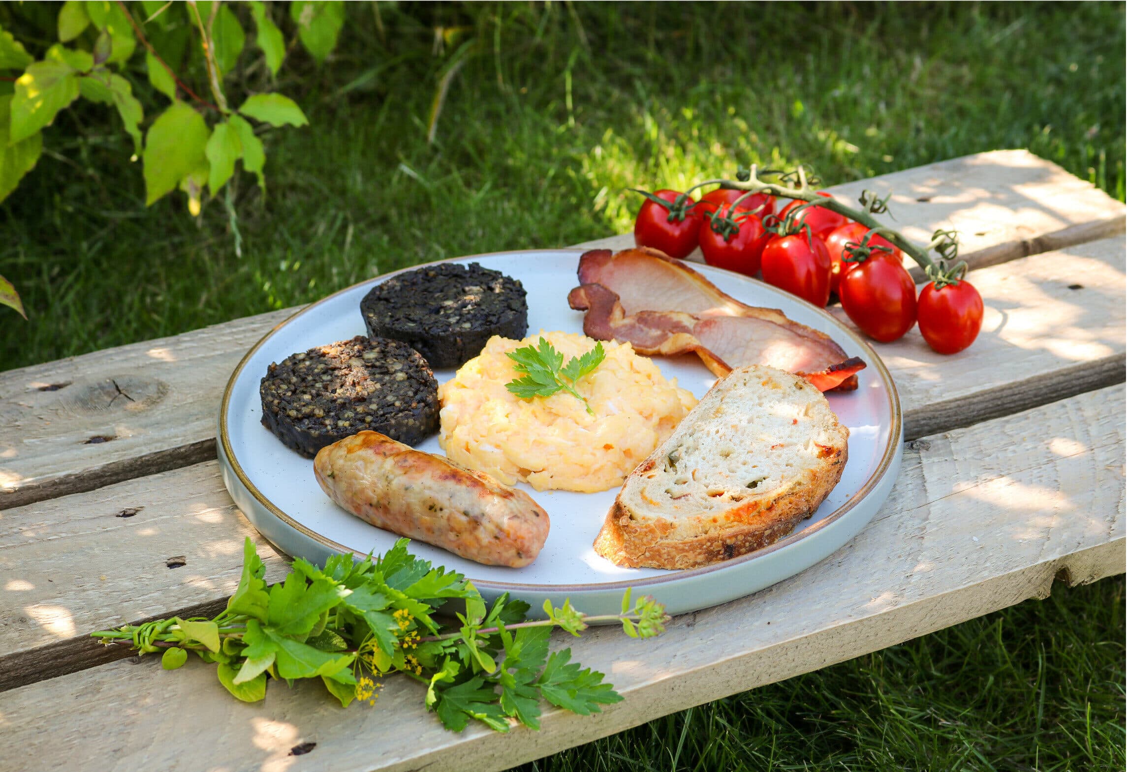 Plate of black pudding, sausage, bacon, scrambled eggs, tomatoes and bread on a wooden bench
